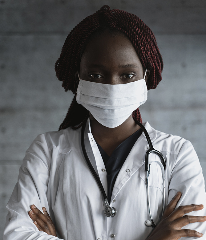 a black woman-presenting person in white scrubs wearing a white mask and stethoscope around their neck and looking powerfully into the camera