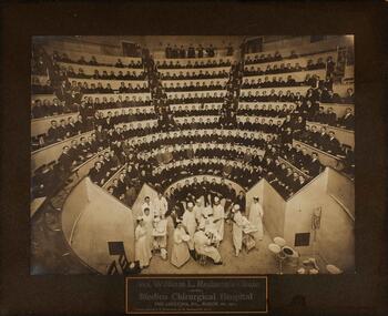 a full surgical theater in 1902 filled with white men in suits, 2 white women nurses assist the surgeons on the floor
