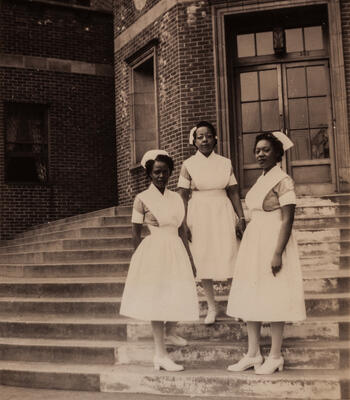three black women nurses stand on the steps outside a hospital building 