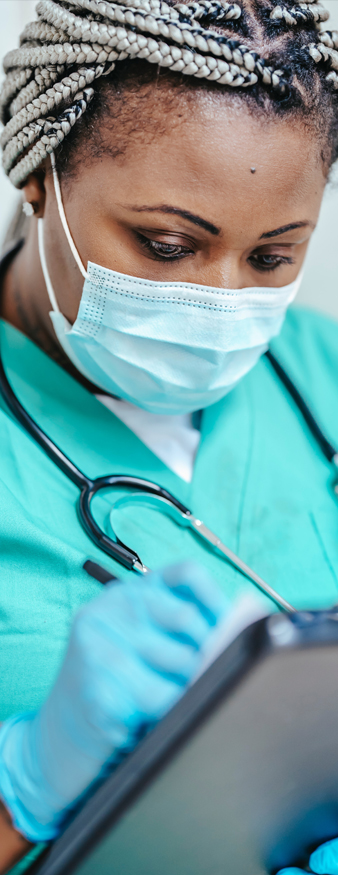 close up of a black woman-presenting doctor wearing blue scrubs and a face mask writing on a clipboard
